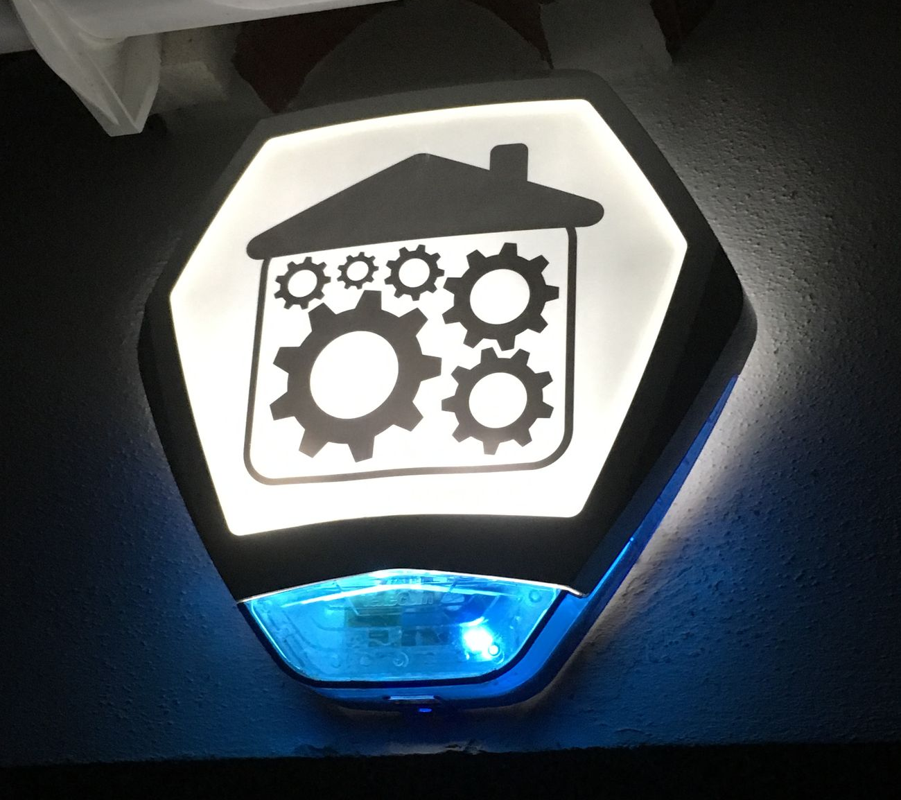 Illuminated sign: a house with gears inside, mounted on a wall with a blue light at the bottom.