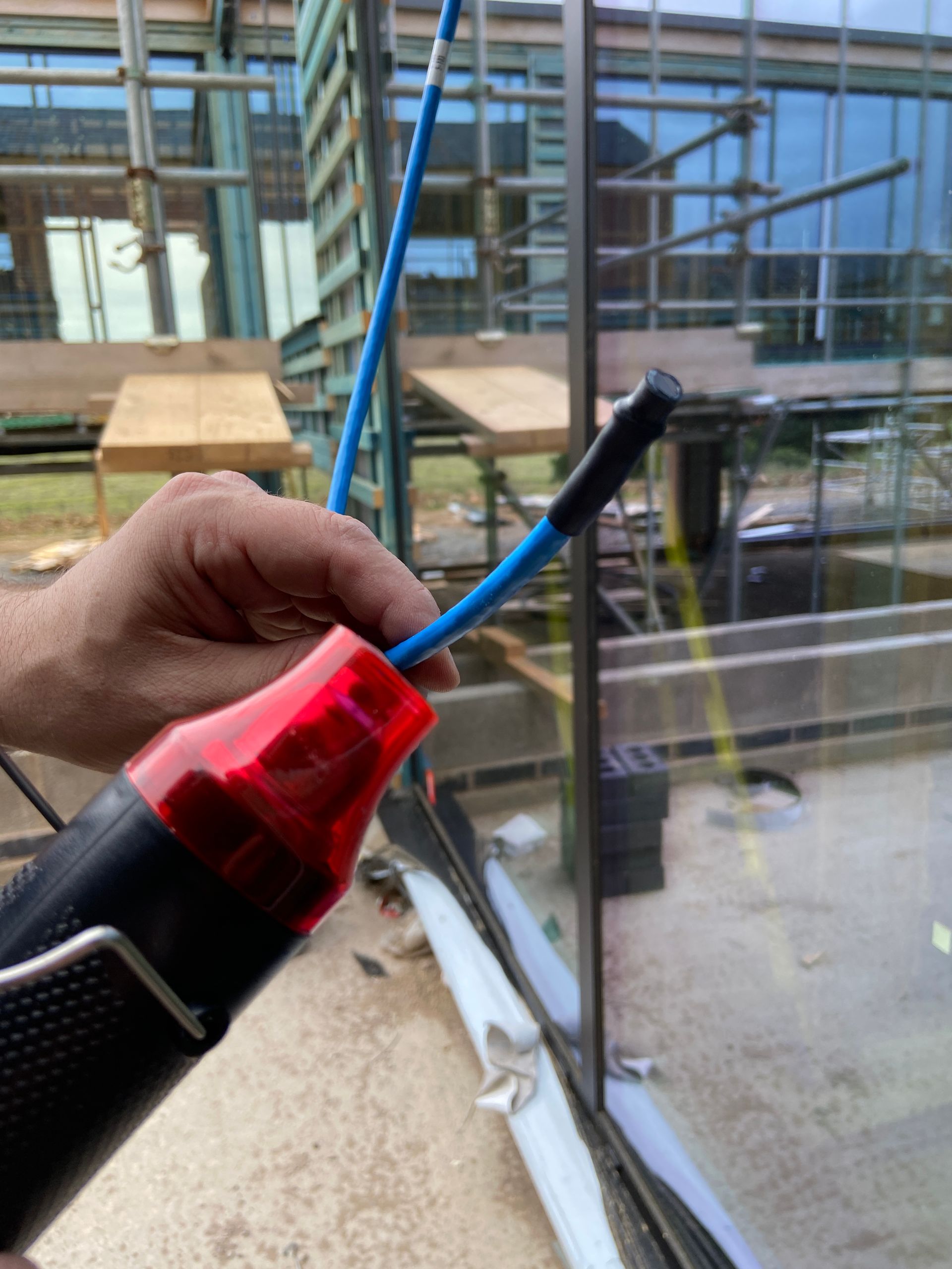 A hand using a heat gun on a blue cable near a window. Building construction visible in the background.