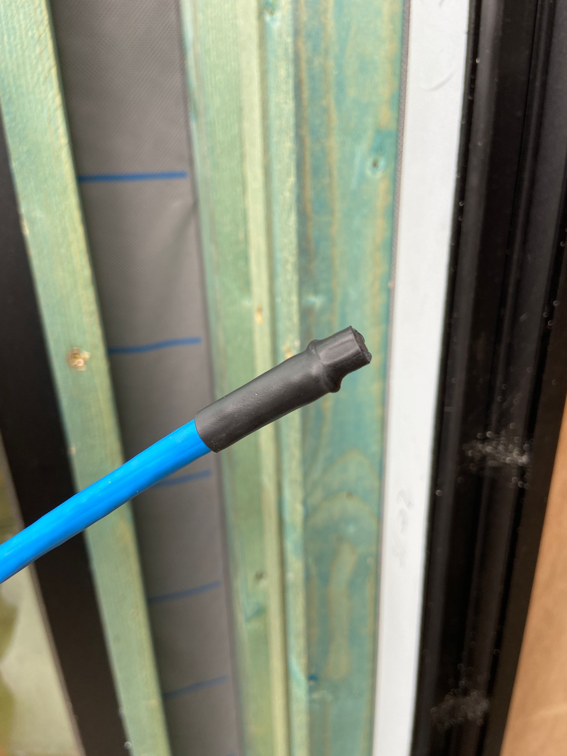 Blue wire with black, rubber-like tip, held against a wooden frame with blue-green vertical boards.