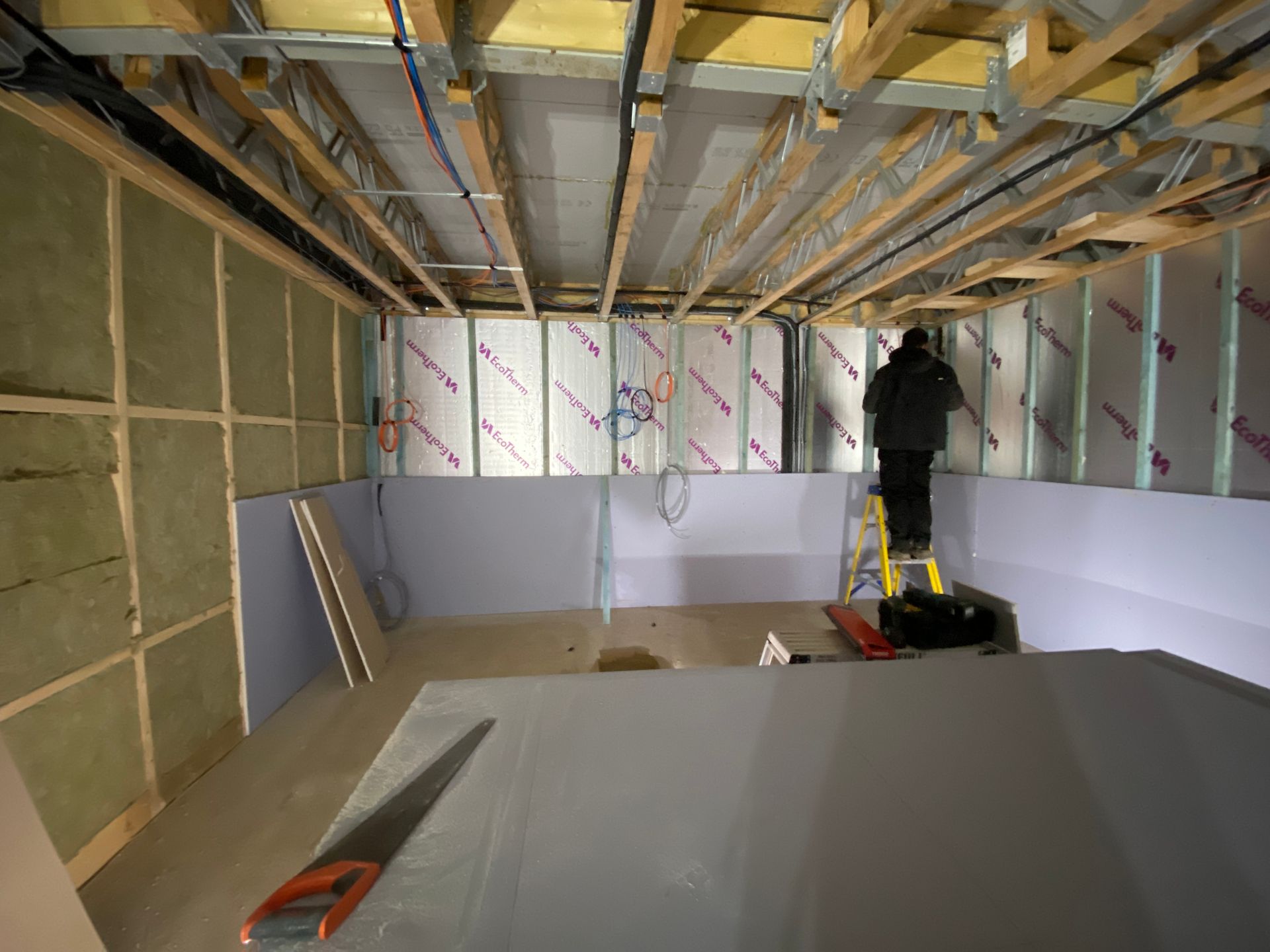 Man on a ladder in a room under construction, with exposed beams, insulation, and drywall.