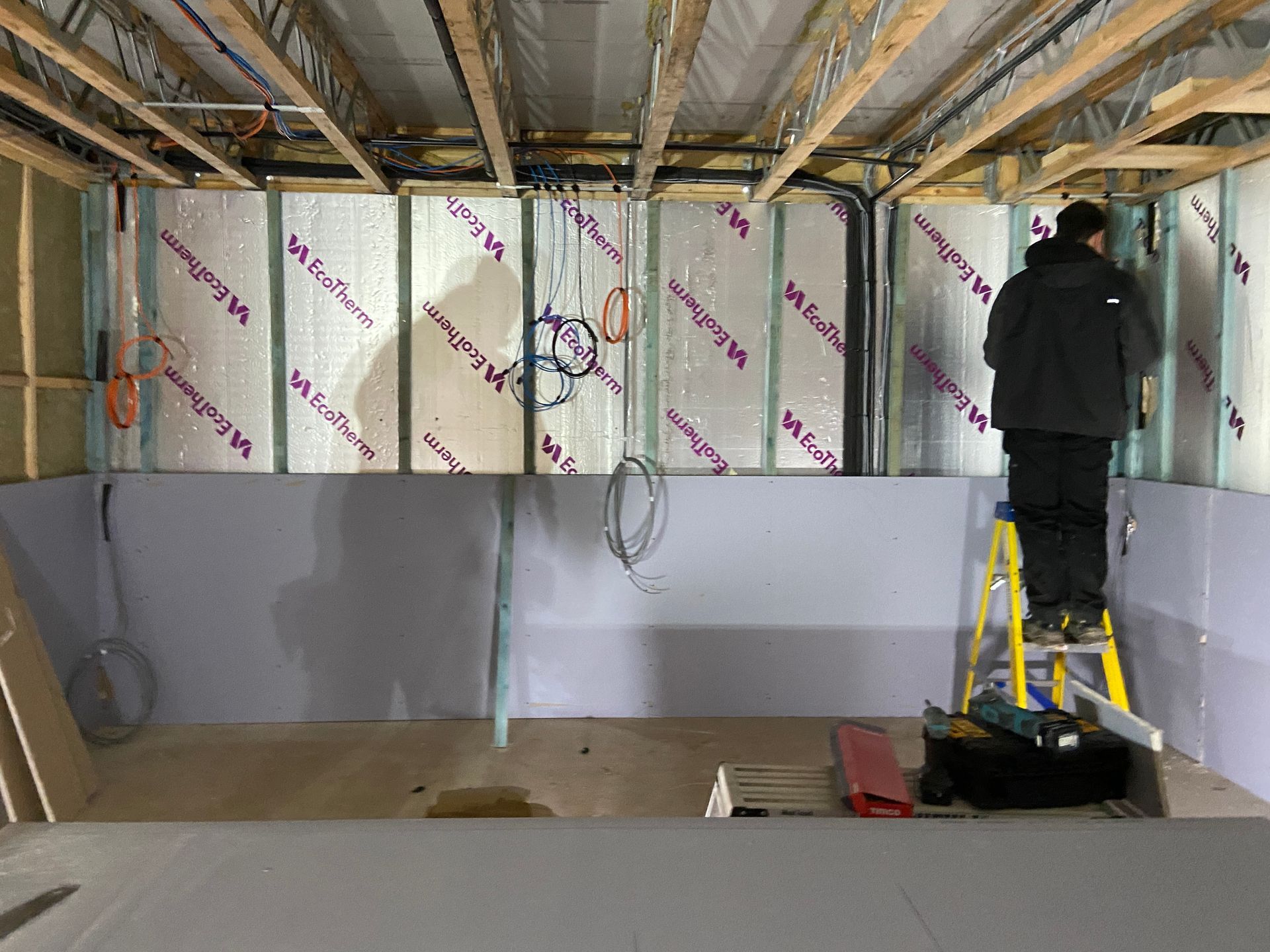 Man on a ladder installing drywall in a room under construction, insulation visible.
