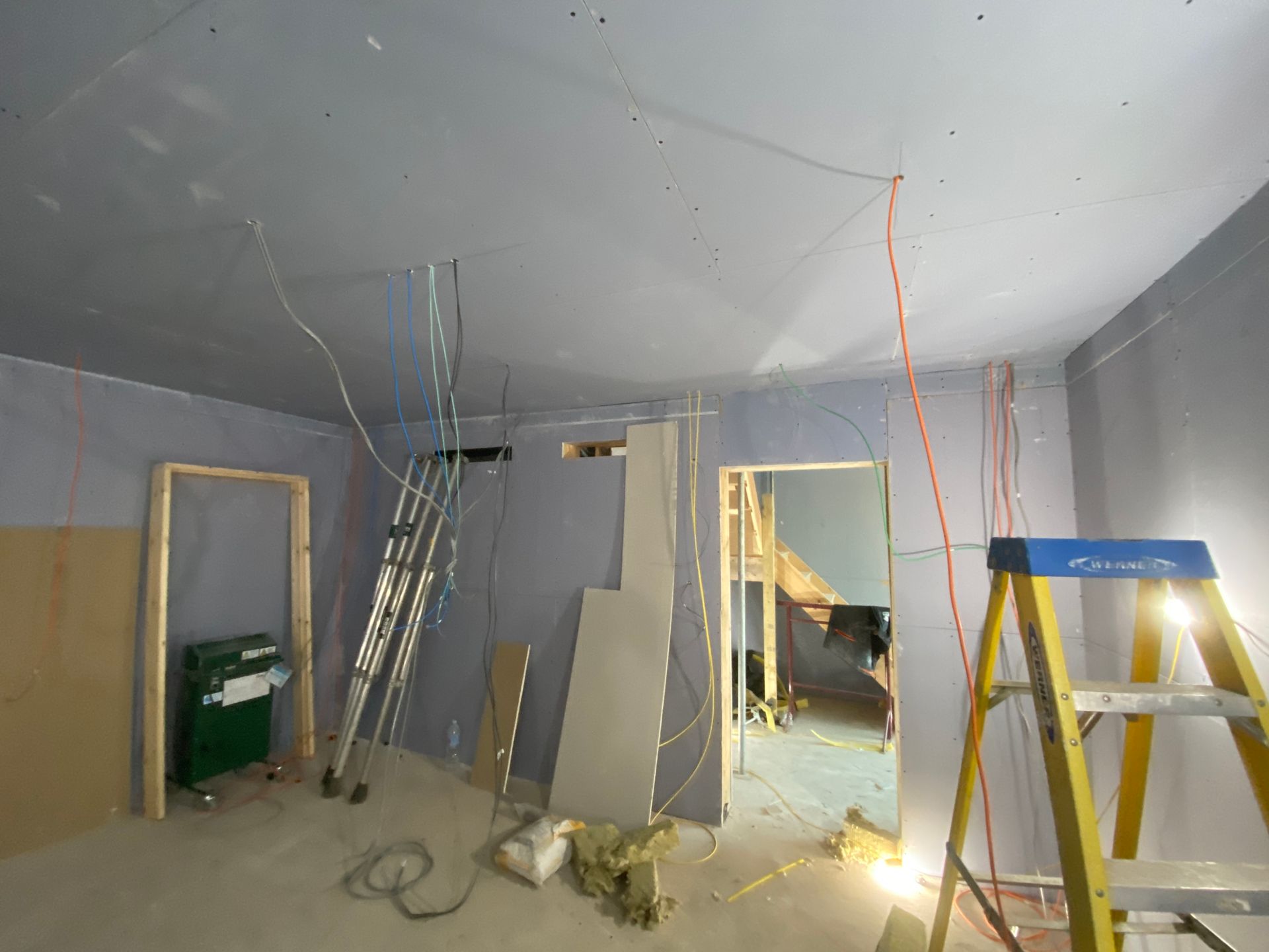 Interior room under construction, with drywall and exposed wiring. Ladder and building materials visible.