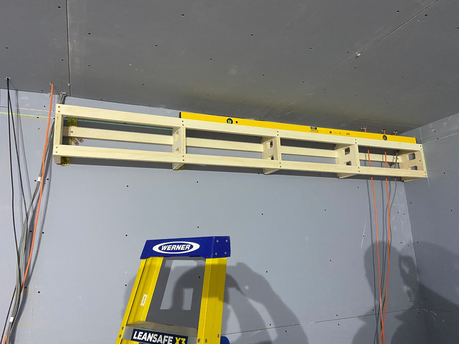 Wooden shelf being installed on a gray wall with a yellow ladder in front. A level sits on the shelf.