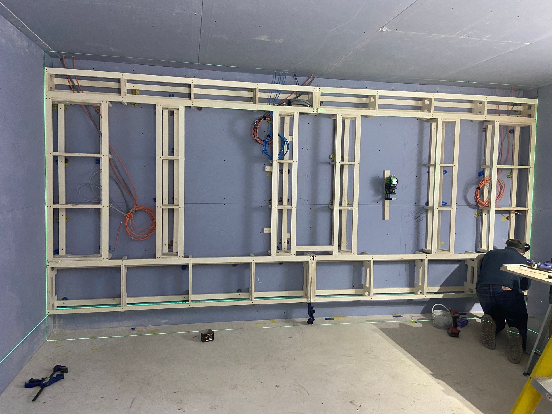 Wooden framework construction against a blue wall; a person works on the lower right. Electrical wiring visible.