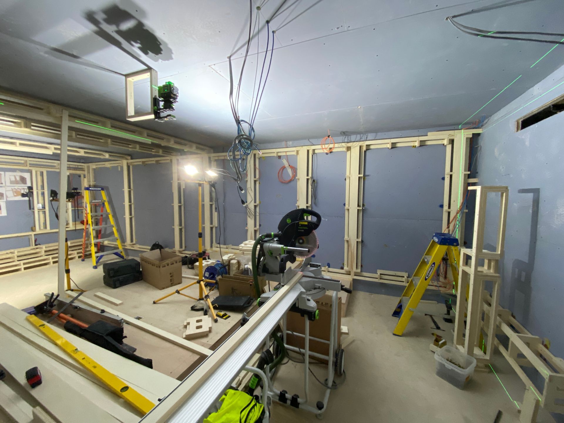 Interior construction site, wooden frames, tools, ladders, exposed wiring, and blue walls.