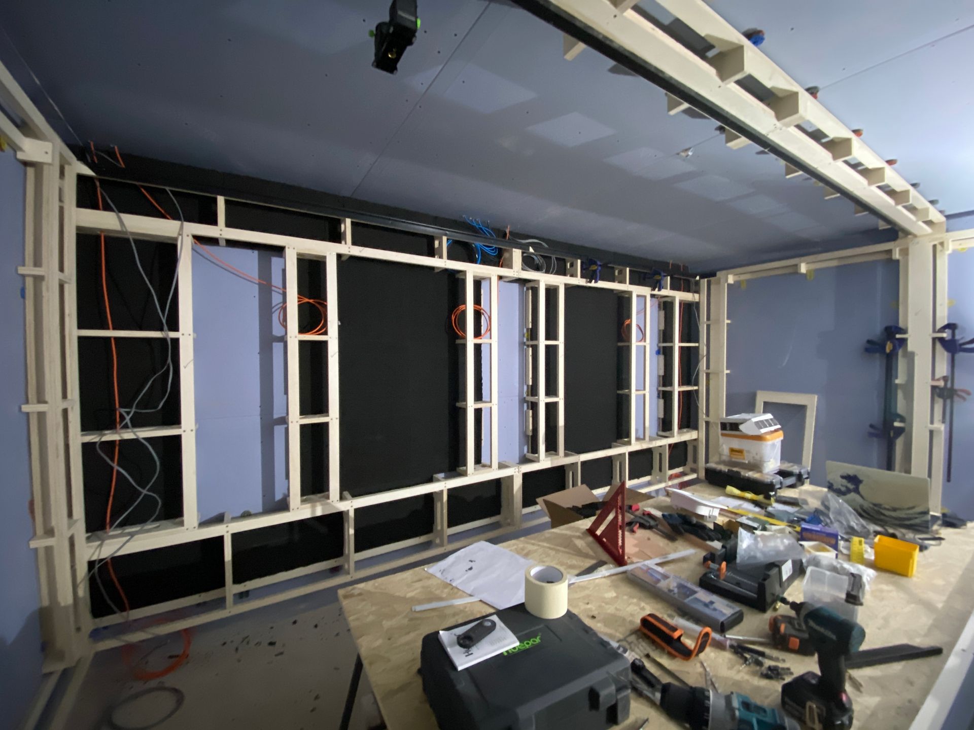 Interior room under construction, with wooden frame, wiring, black panels, and tools on a workbench.