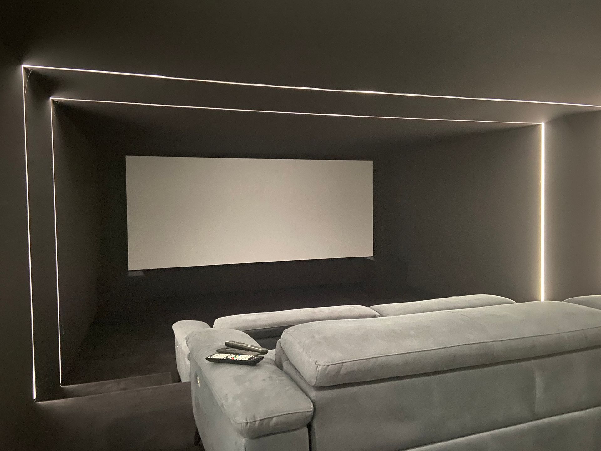 Modern home theater with gray walls, white screen, and gray sofa, illuminated by linear lights.