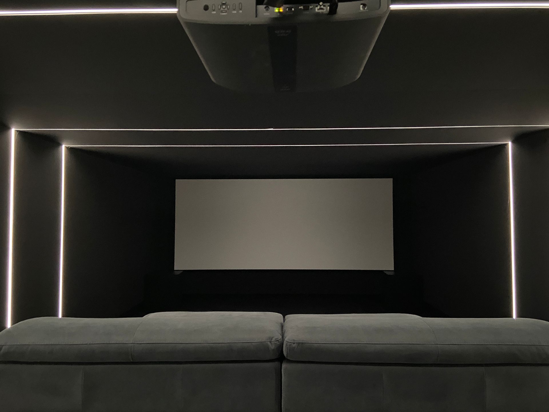 Dark home theater with gray couch, projector, and screen, illuminated by neon lights.