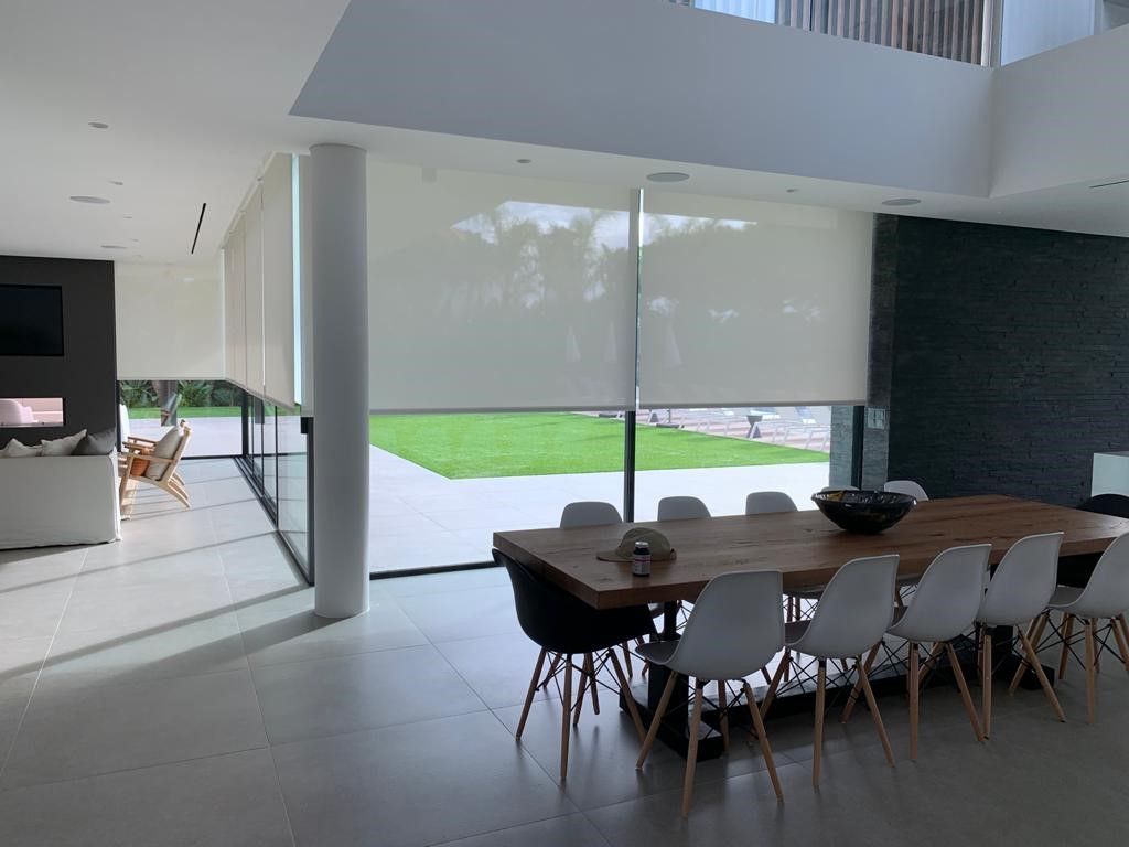 Modern dining room with large windows, white blinds, wooden table, and white chairs.
