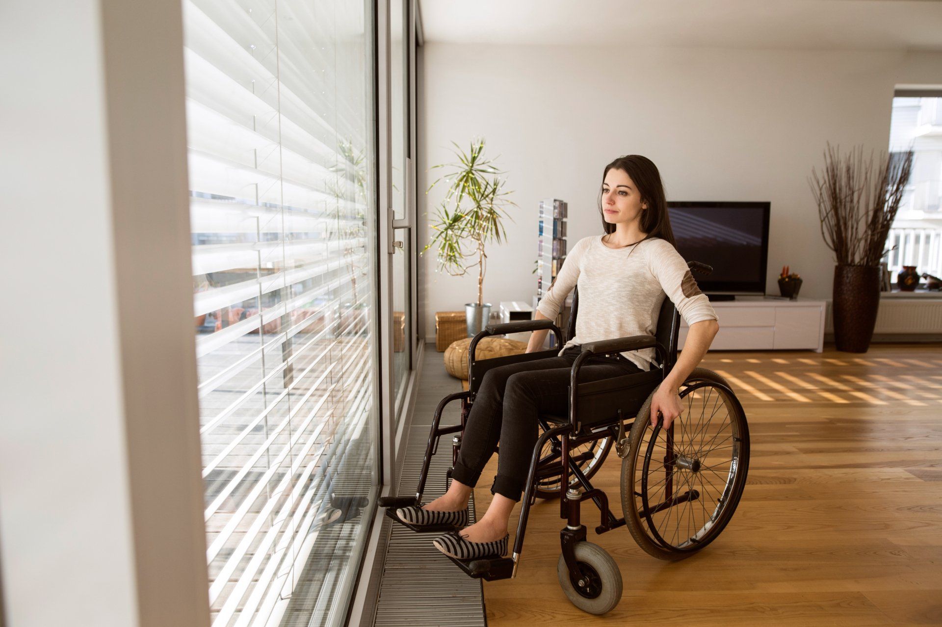Woman in wheelchair looking out large window, wooden floor, neutral tones, sunny interior.