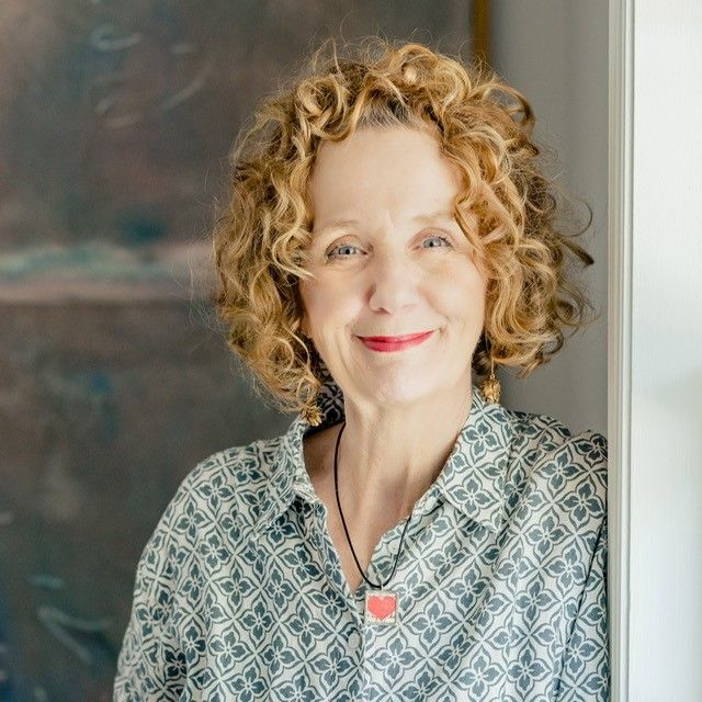 A woman with curly hair is leaning against a wall and smiling