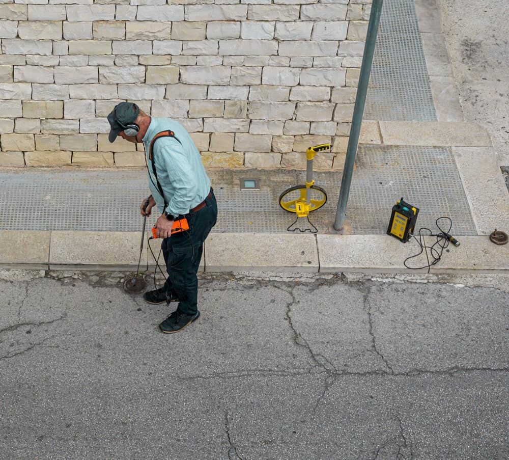A Man Wearing Headphones Is Using A Metal Detector On The Sidewalk — DAVMAC Plumbing in Wauchope, NSW