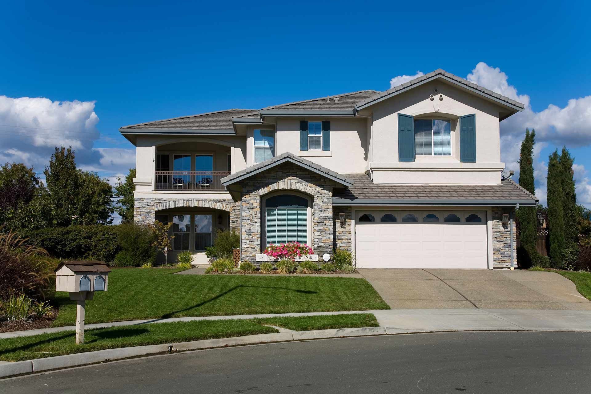 Two-story beige house with a gray roof and stone accents, driveway, and a mailbox under a blue sky.
