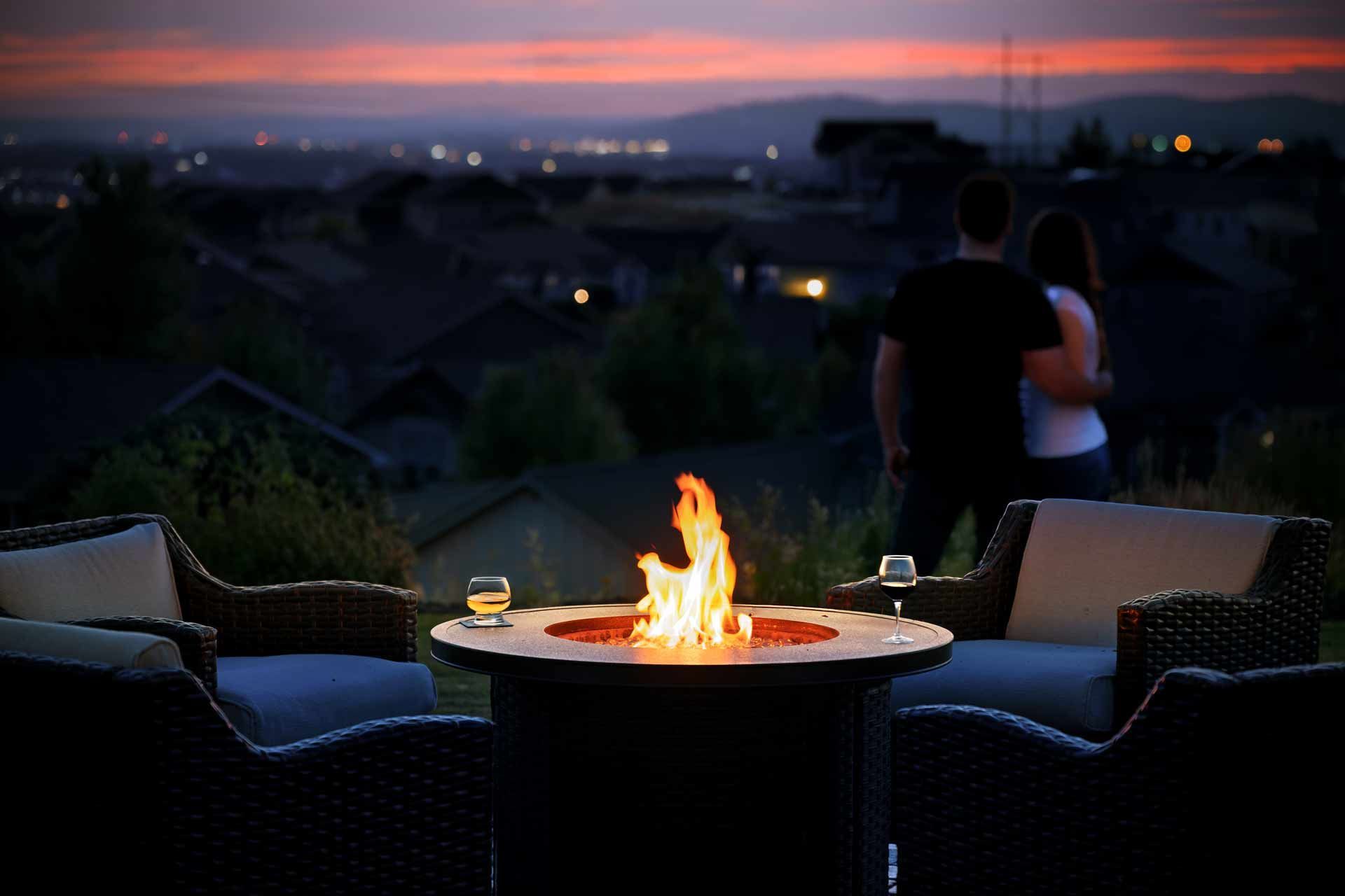 Couple gazing at sunset over town, by lit fire pit with wine glasses.