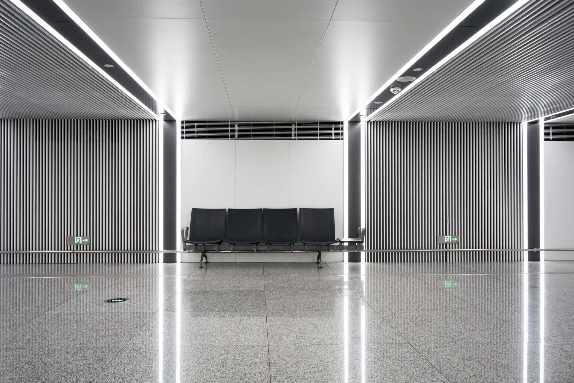 Modern airport waiting area with gray bench, vertical ribbed walls, and bright ceiling lights.