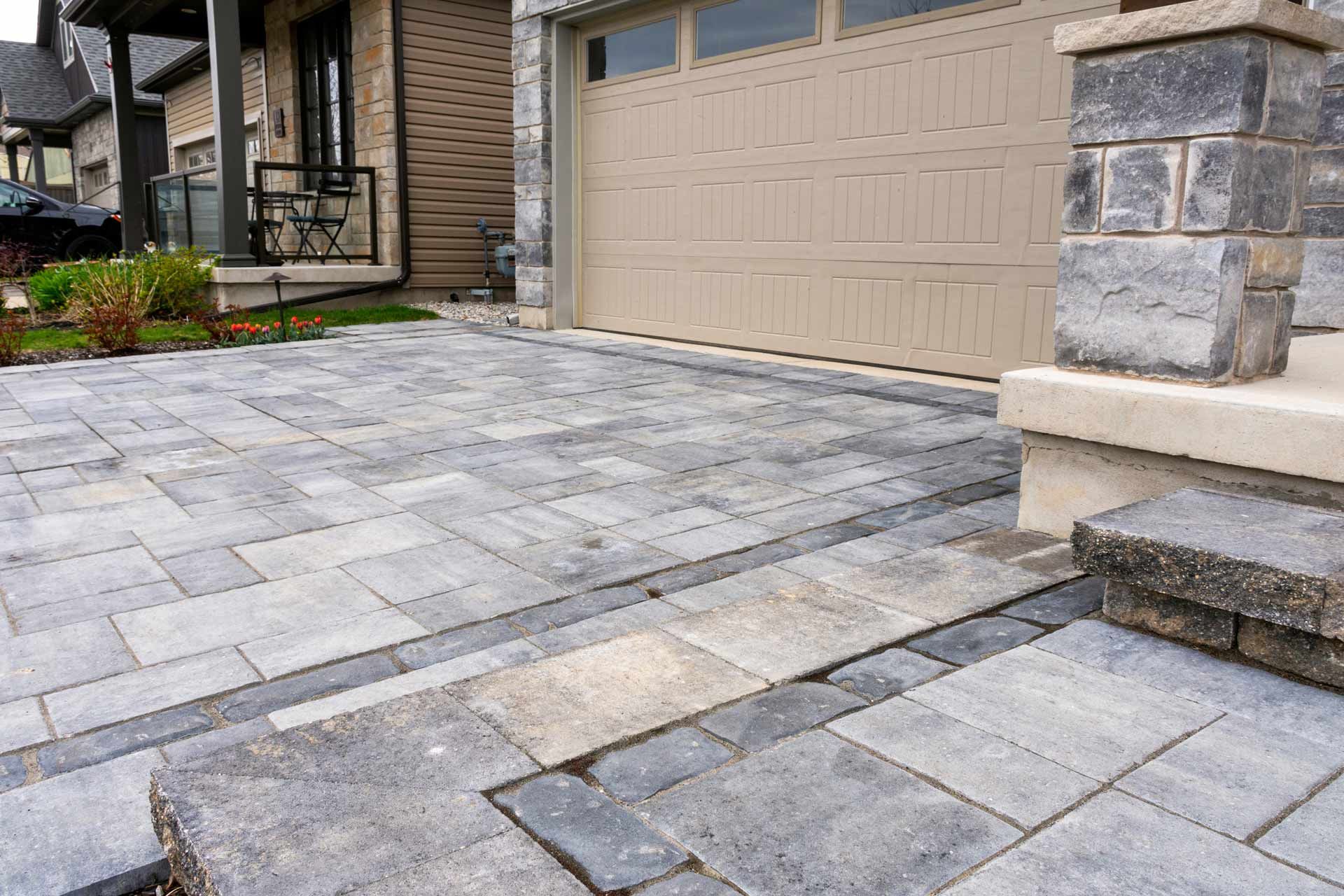 Paver driveway and walkway leading to a beige garage, next to a house with a black railing.