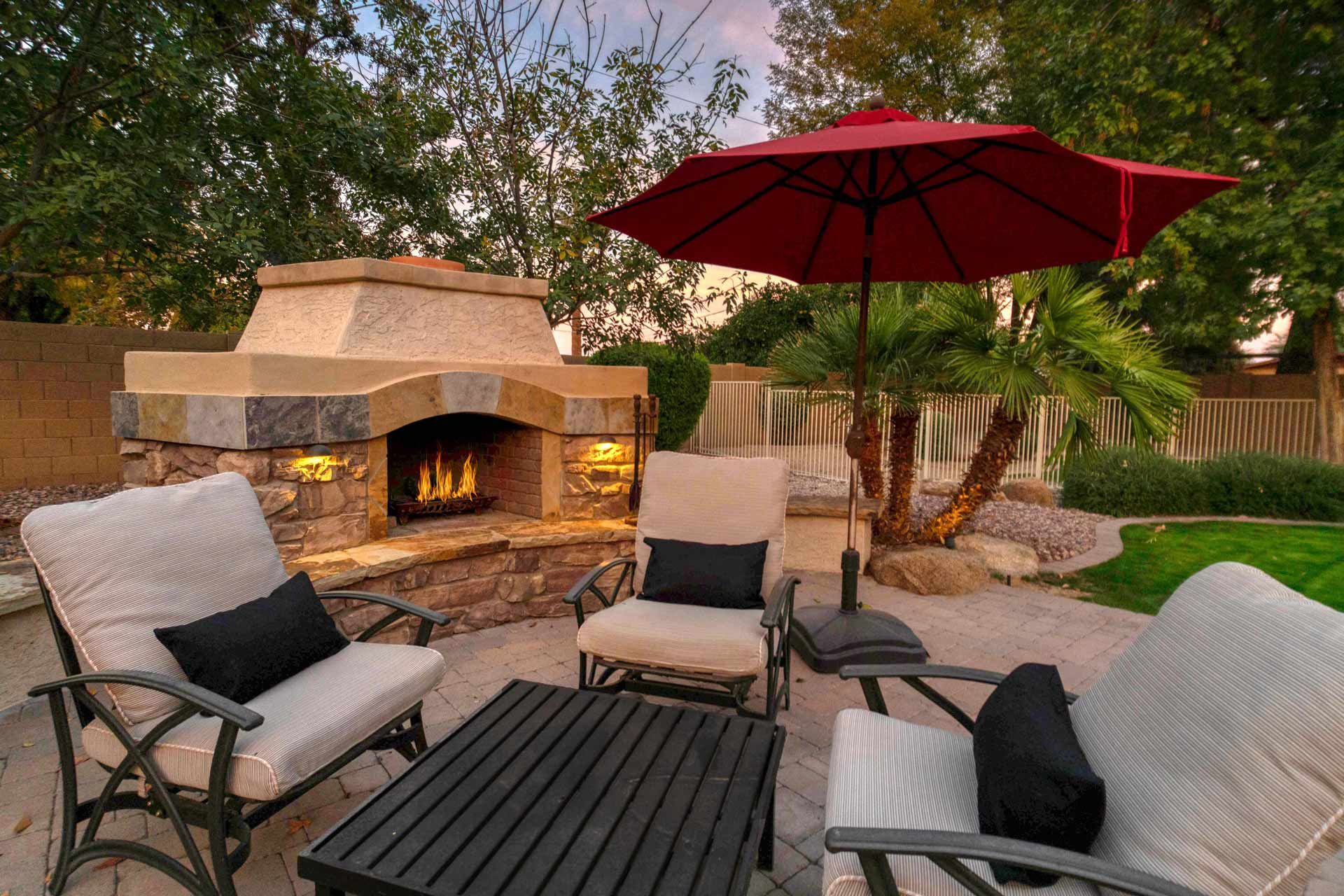 Outdoor patio with fireplace, seating, red umbrella, and palm tree.