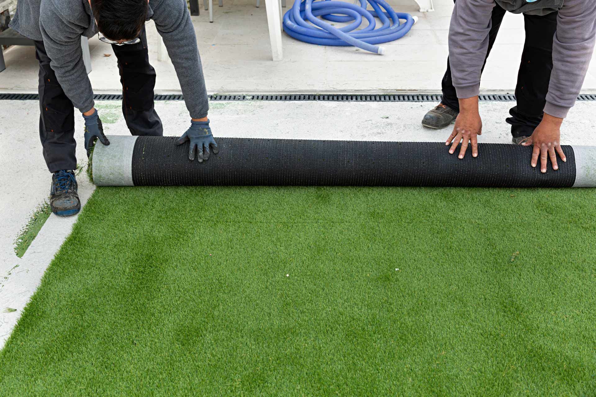 Two people unrolling a roll of green artificial turf on a concrete surface.