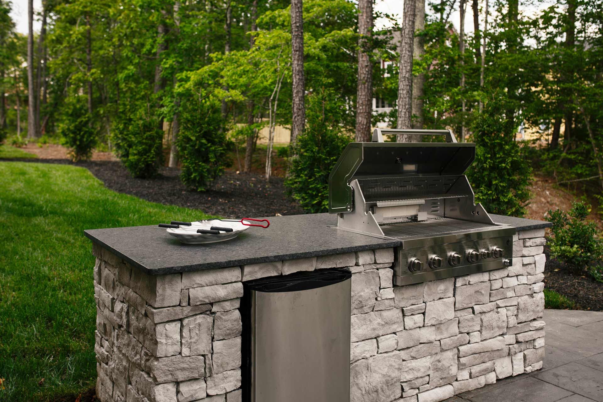 Outdoor kitchen with a stone facade, a grill, counter, and a trash bin. Green grass and trees in the background.