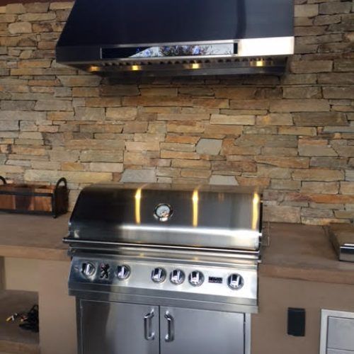 Outdoor stainless steel grill under a vent hood, set against a stone wall and concrete counter.