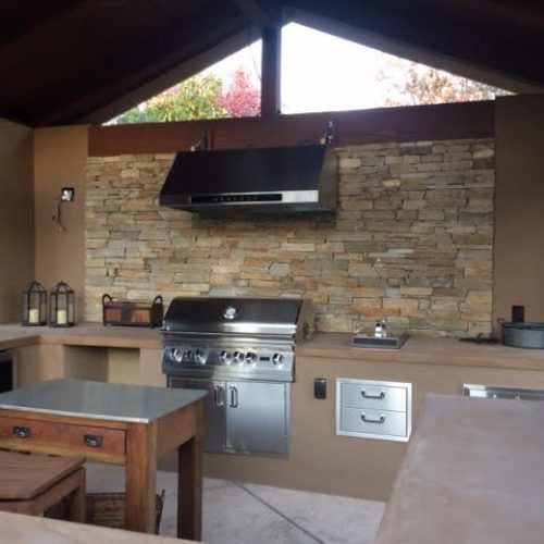 Outdoor kitchen with grill, stone wall, stainless steel hood and cabinets. Brown and neutral tones.