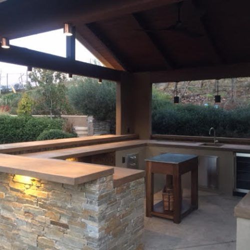 Outdoor kitchen with stone accents, bar, sink, and grilling area. Brown wooden overhead beams and lighting.