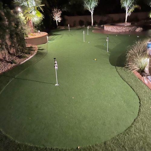 A well-lit backyard putting green with several golf flags. Artificial turf surrounds a pool and landscaping.