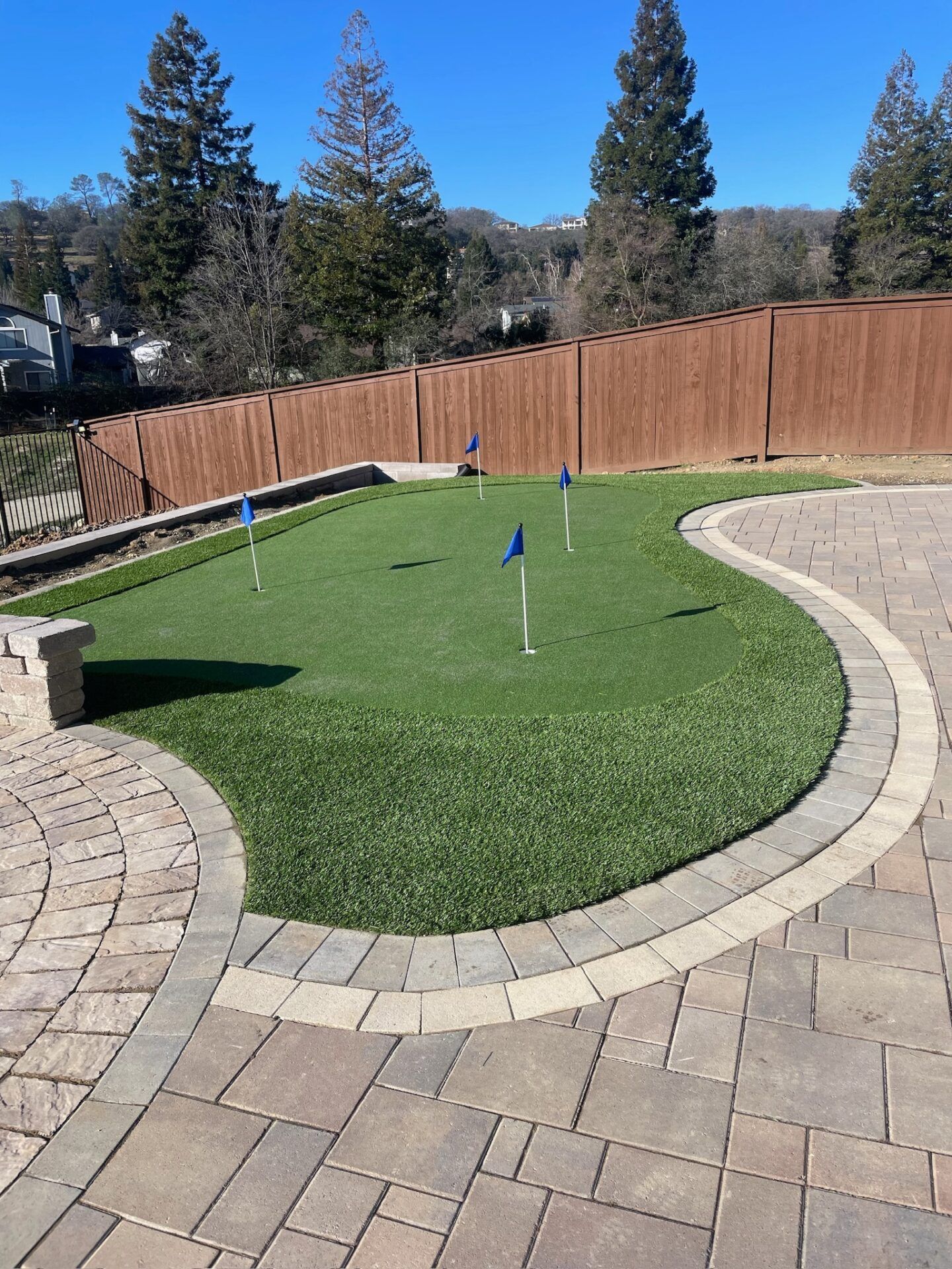 Backyard putting green with blue flags, surrounded by stone patio and wooden fence.