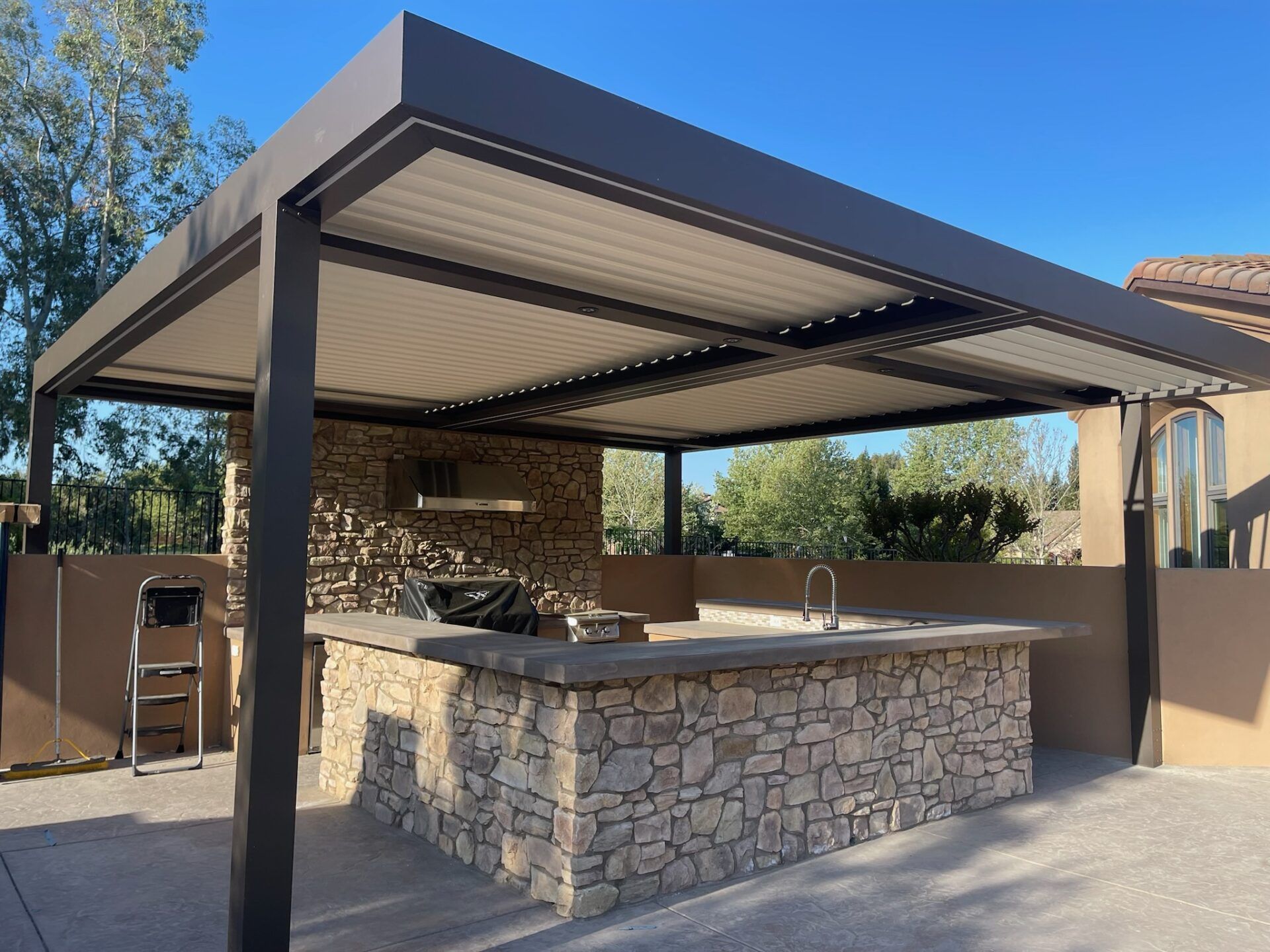 Outdoor kitchen with stone facade, under a dark pergola with adjustable roof. Sink, grill, and counter.