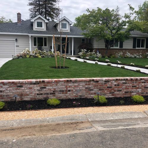 Gray house with brick wall, green lawn, white flowers, and walkway.
