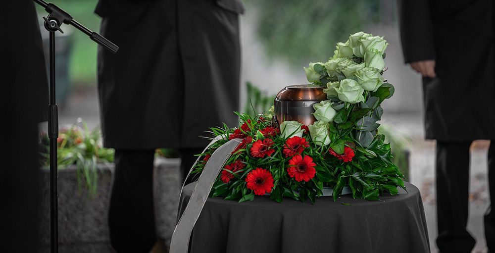 A funeral ceremony with a floral arrangement and urn on a table, people standing.
