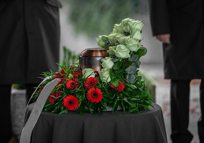 Bronze urn with floral arrangement at a funeral. Two figures in black coats stand nearby.