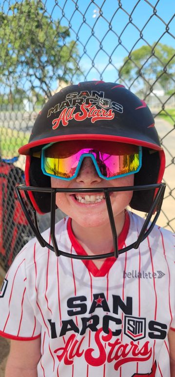 A person wearing a hat and orange shirt, holding a baseball bat on their shoulder on a baseball field.