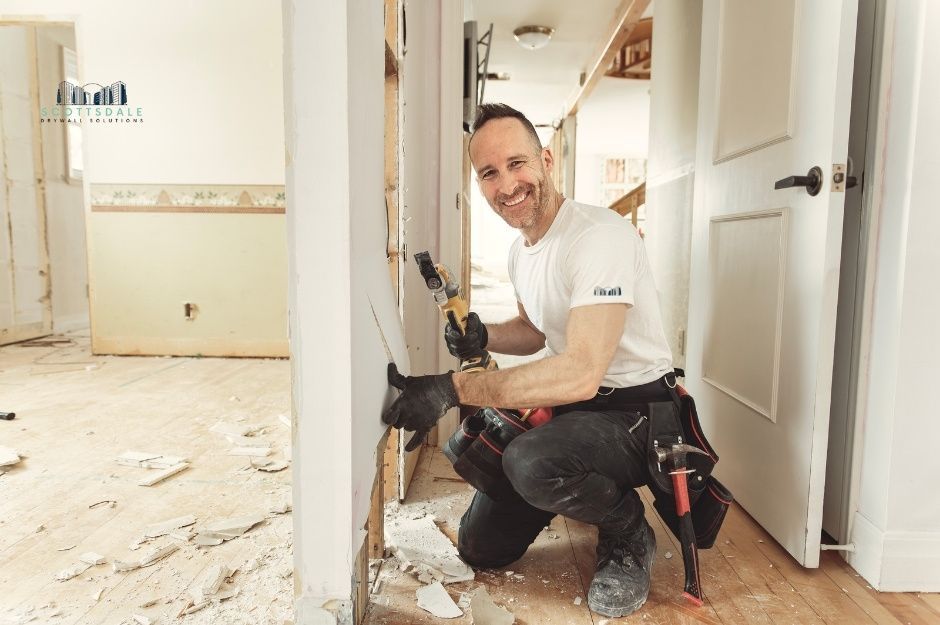 A drywall repair company professional kneeling while working on wall repair, holding a drill tool in a residential hallway with white walls and flooring materials scattered around near N Pinion Hills Ct, and W Wolf Run Dr, Anthem, AZ.