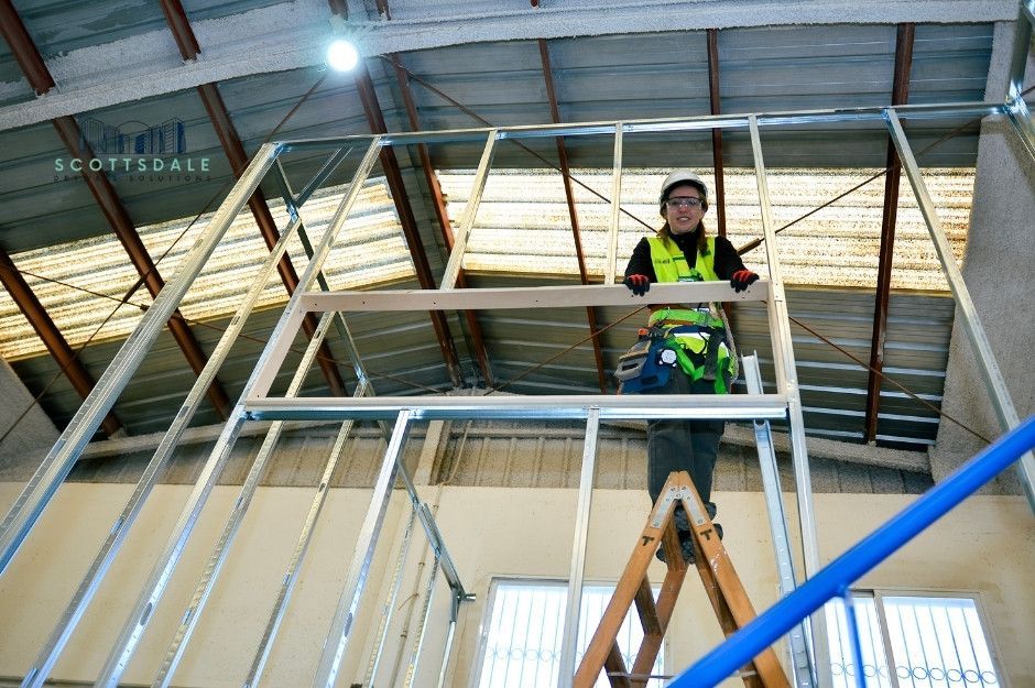 A drywall repair company worker in a high-visibility vest stands on metal scaffolding inside a residential construction site. She is inspecting the ceiling framework. The scaffolding has safety railings and shows exposed structural beams near S Warner Dr, and W Hawks Eye Ave, Apache Junction, AZ.