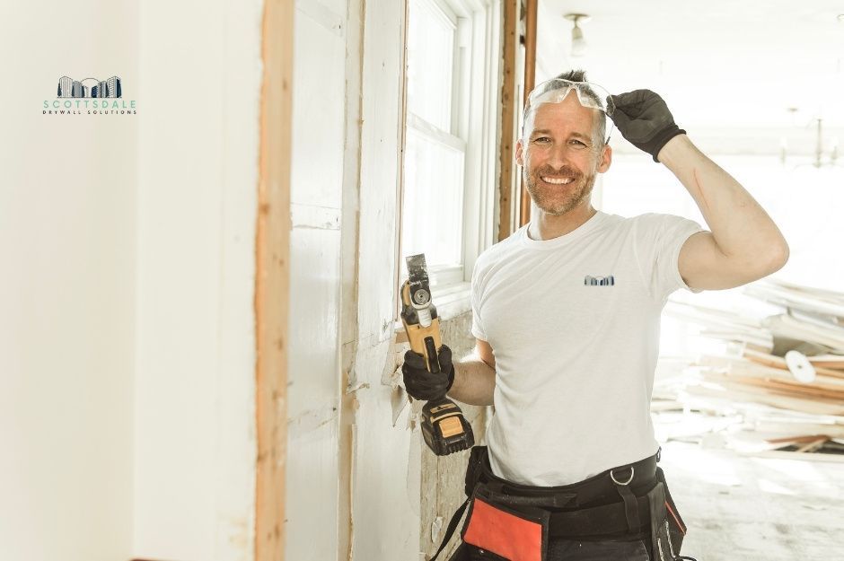 A drywall repair company worker smiling in a white shirt and a tool belt, holds a power drill while standing next to white drywall panels in a bright residential construction space near Valley Cir, and Valley Dr, Black Canyon City, AZ.