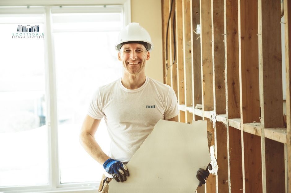 A drywall repair company worker in a white hard hat and shirt smiles while holding a drywall sheet against wooden wall studs in a bright residential construction site. Professional contractor prepares for installation work near W Casa Mirage Ct, and W Mirage Loop, Casa Grande, AZ.