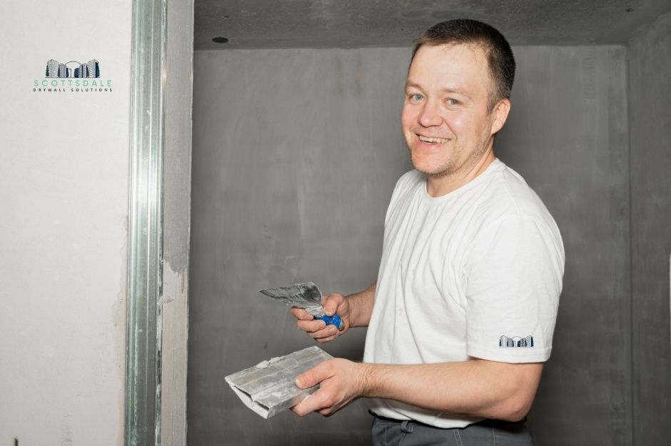 A drywall repair company technician smiles while holding a putty knife and applying joint compound to repair drywall in a residential construction site with exposed framing. The worker wears a white t-shirt, smoothing the compound on a wall surface near Dewey Ave, and S 1st St, Coolidge, AZ.