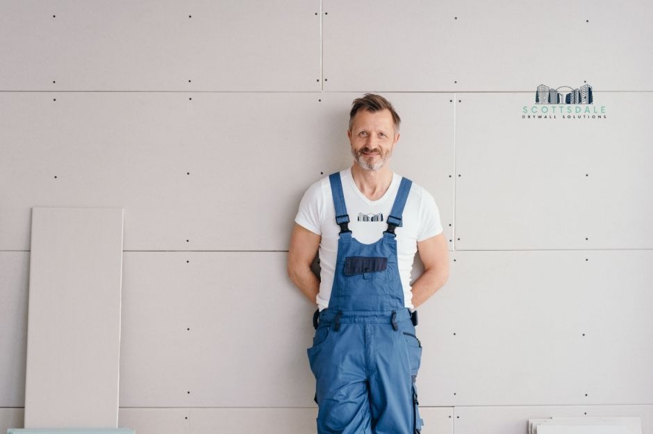 A drywall repair company professional stands confidently against a white drywall wall. He wears blue overalls over a white t-shirt with his hands behind his back, ready to tackle any residential drywall project near E Longhorn Ln, and S Maverick Trail, Cordes Lakes, AZ.
