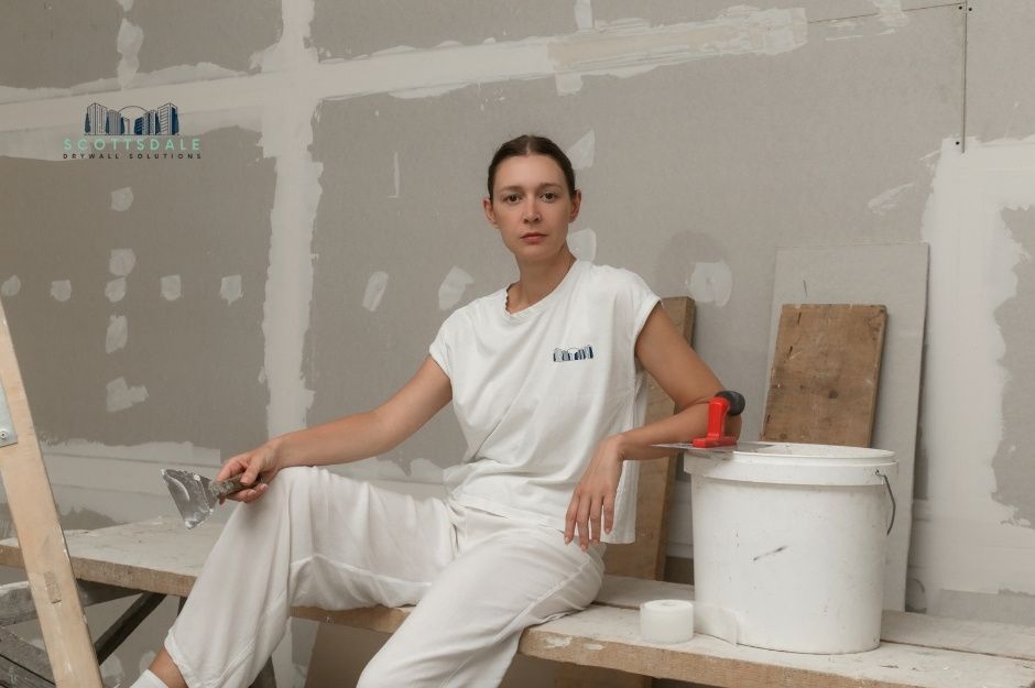A drywall repair company professional woman in white work clothes sits in a room with freshly mudded drywall walls. She holds a putty knife tool with a white bucket and painting supplies nearby during residential renovation work near E Phoenix Ave, and N Sunshine Blvd, Eloy, AZ.