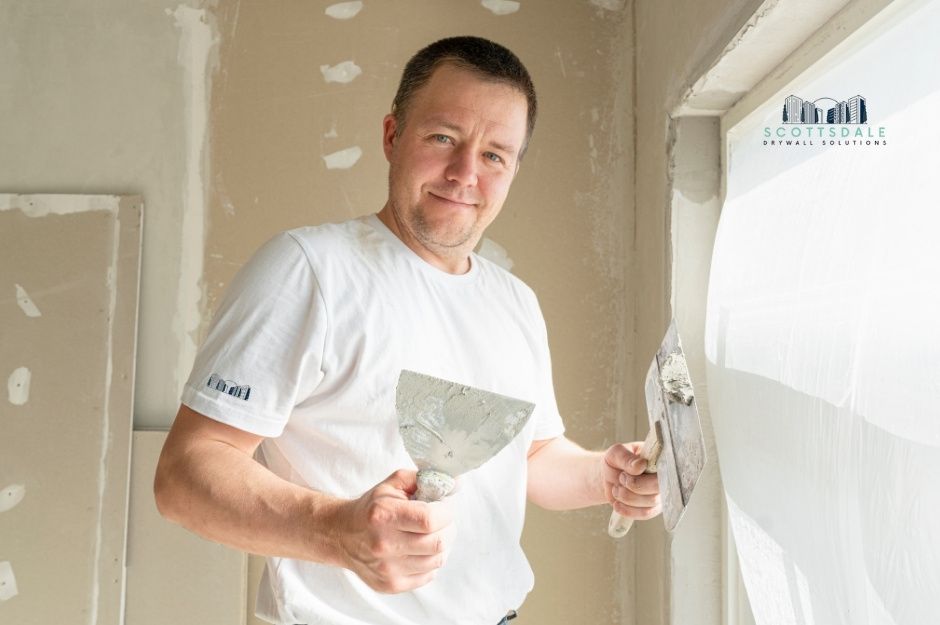 A drywall repair company professional smiling while holding a putty knife and joint compound, wearing a white t-shirt, standing in a bright interior of a commercial construction site with white walls and natural lighting from windows near N Crestwood Ct, and W Rushmore Way, Florence, AZ.