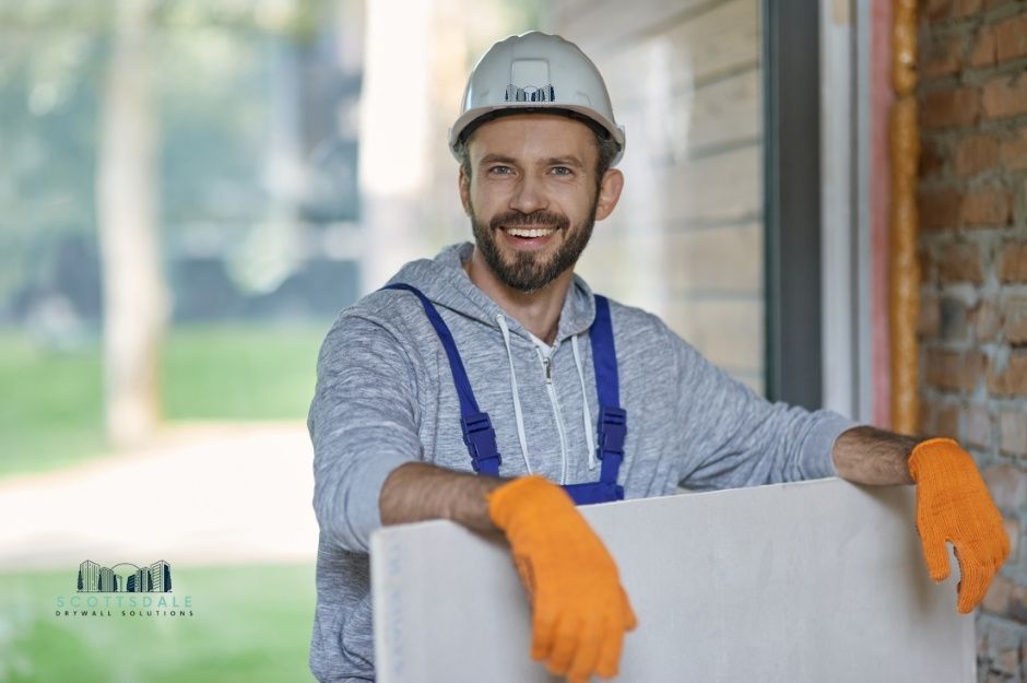 A drywall repair company worker in a hard hat, and orange gloves is holding a white drywall sheet. He has a beard, smiling at the camera, and is at a residential construction site near Cll Sahuaro, and E Calle Barbarita, Guadalupe, AZ.