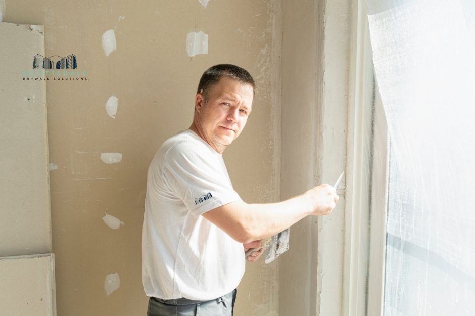 A drywall repair company professional in a white shirt applies joint compound to a patched drywall section using a putty knife, smoothing the repair work on a commercial construction site near W Rainwater Dr, and S 54th Ave, Laveen, AZ.