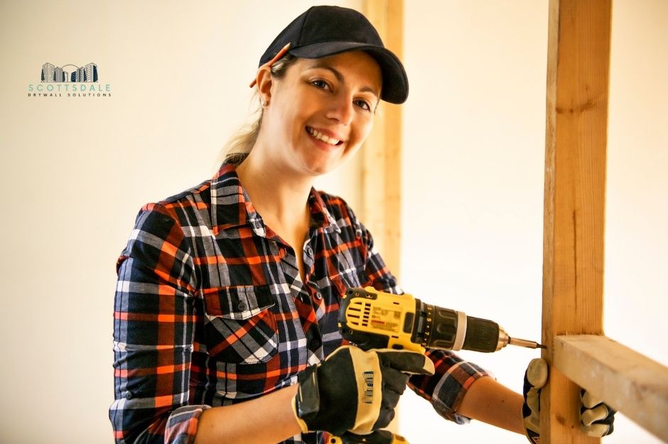 A drywall repair company woman, smiling in a black cap and plaid flannel shirt, holds a yellow cordless drill while working on residential interior wall repairs. She is confident and skilled at her craft near W Sunland Ave, and S 26th Ln, Liberty, AZ.