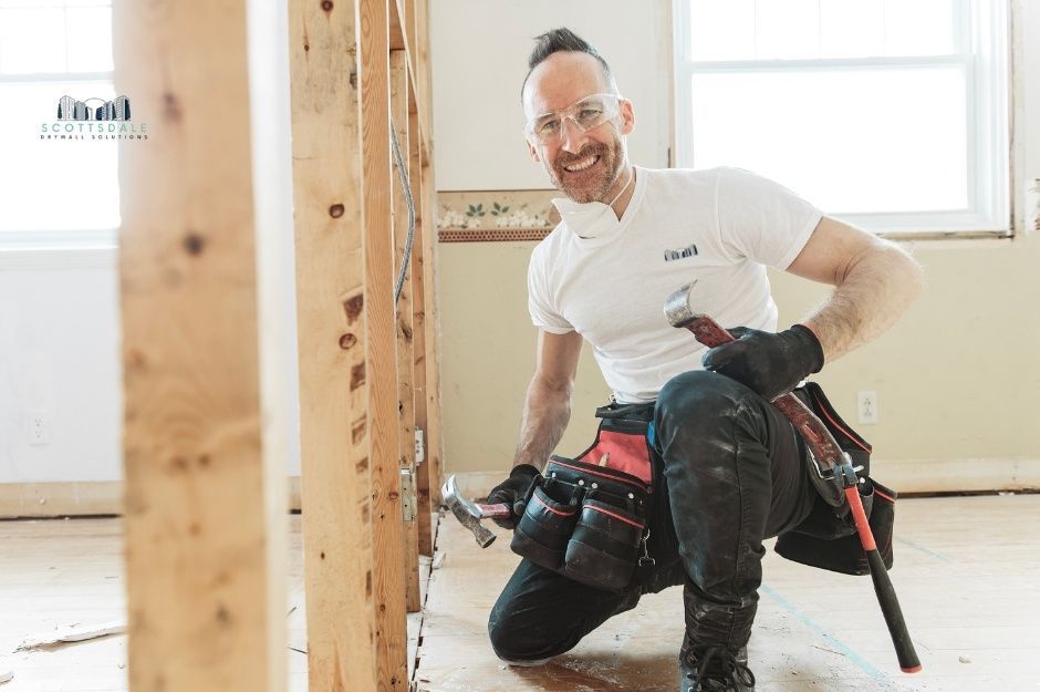 A drywall repair company worker kneels on the floor in a white shirt, holding a hammer while working on wooden wall framing in a bright, unfinished commercial construction site with exposed studs and a window visible in the background near E Scooter Ln, and S Ivory Ln, Mayer, AZ.