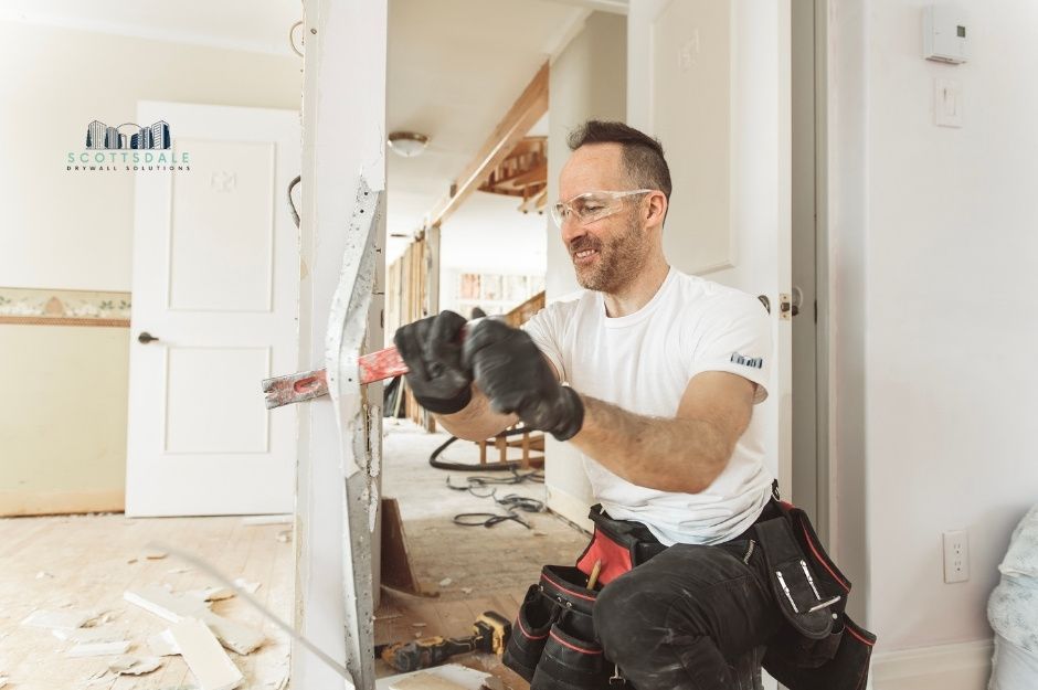 A drywall repair company worker in a white t-shirt and glasses uses a panel lifter on interior walls. He is working with drywall materials and equipment in a residential construction setting near E Mesquite St, and E Desert Hills Dr, New River, AZ.