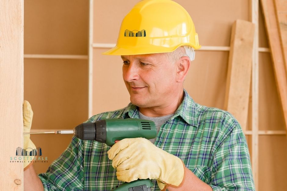 A drywall repair company worker, wearing a yellow hard hat and a green plaid shirt, holds a power drill while working on a drywall installation. He is focused on his task with residential wooden framing visible in the background near N Superstition Ln, and E Starlight Way, Paradise Valley, AZ.
