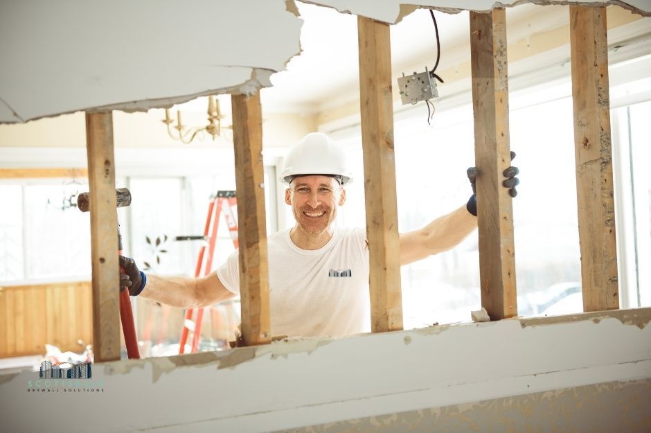 A drywall repair company worker in a white hard hat and shirt stands between exposed wooden wall studs in an unfinished interior space. The professional is positioned among the framing during residential construction work near N Industrial Way, and E Valley Rd, Prescott Valley, AZ.