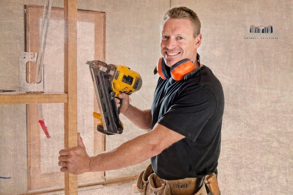 A drywall repair company worker smiles while operating a yellow reciprocating saw on wooden framing in an unfinished room with exposed studs and drywall. He wears a black shirt and tool belt, demonstrating professional residential construction work near E Excelsior Ave, and S 214th St, Queen Creek, AZ.