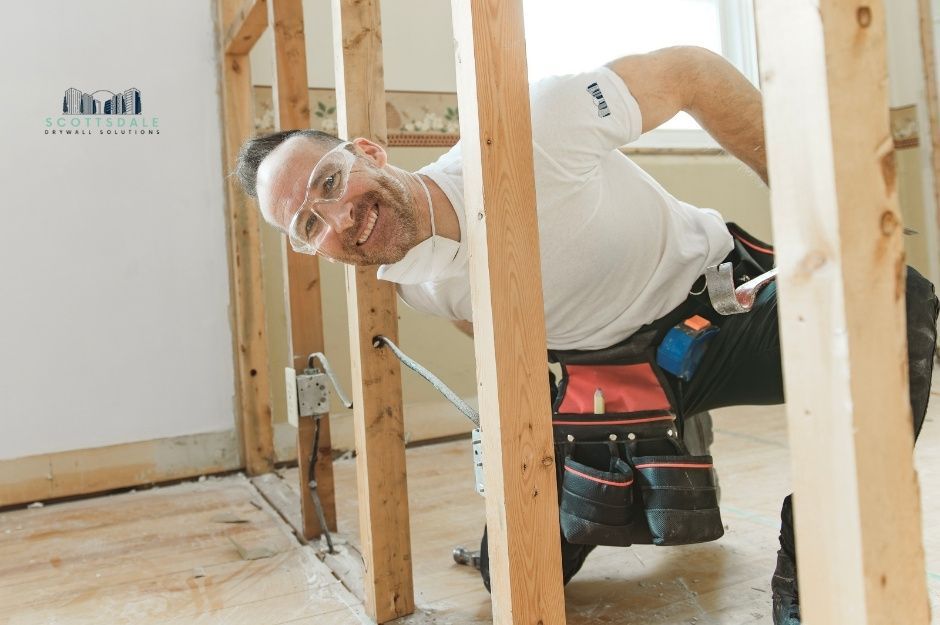 A drywall repair company worker kneels between wooden wall studs in an unfinished residential construction site, reaching toward the framing while wearing a tool belt. He is performing installation and repair work on the exposed wooden framework near Gambol Cir, and Gambol Ln, Spring Valley, AZ.
